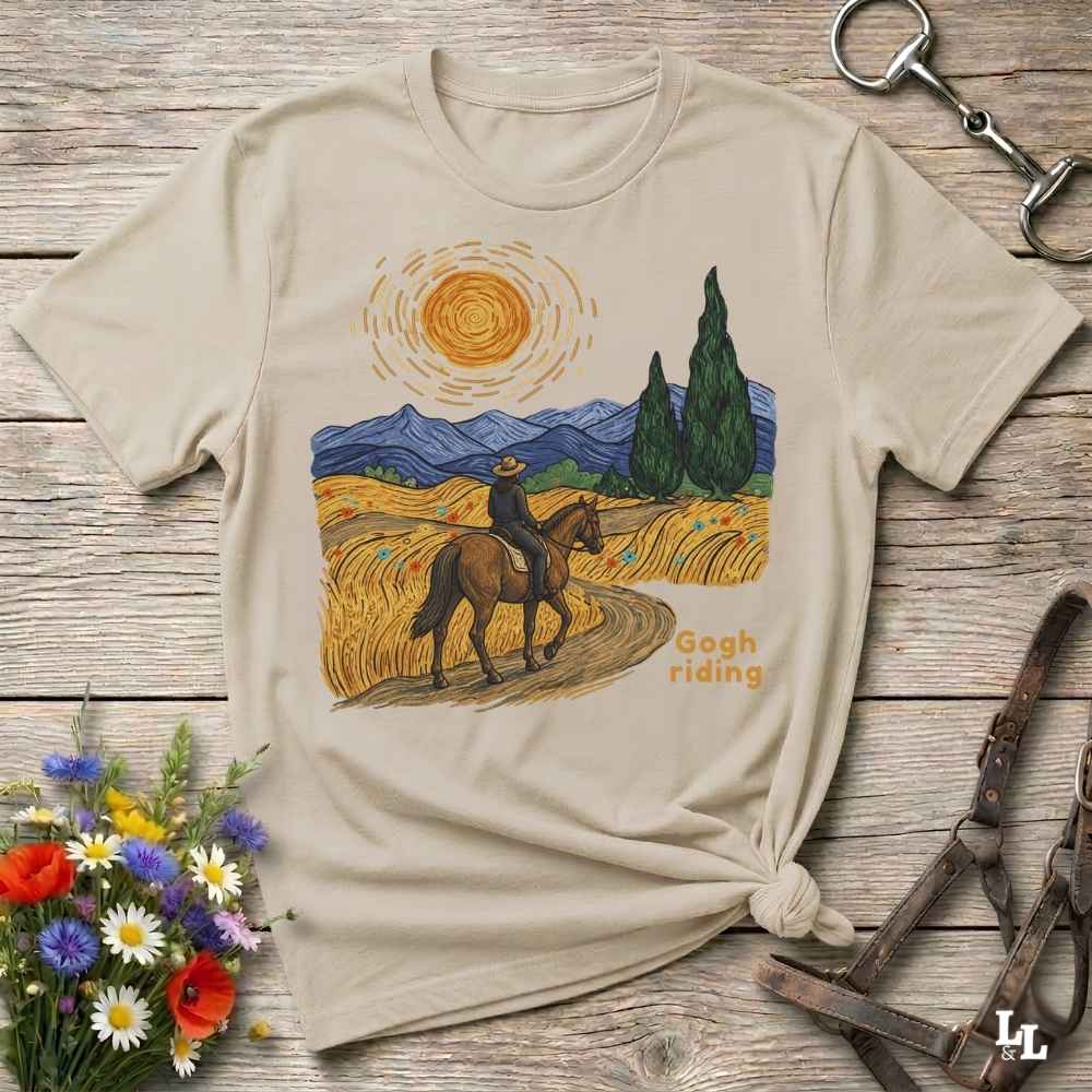 A beige graphic t-shirt laid flat on a rustic wooden surface, styled with equestrian accessories and a small bouquet of wildflowers. The t-shirt features a colorful print in a Van Gogh-inspired style showing a rider on a horse through a golden field under a bright sun, with the text 'Gogh riding'.