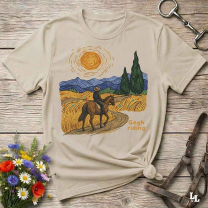 A beige graphic t-shirt laid flat on a rustic wooden surface, styled with equestrian accessories and a small bouquet of wildflowers. The t-shirt features a colorful print in a Van Gogh-inspired style showing a rider on a horse through a golden field under a bright sun, with the text 'Gogh riding'.