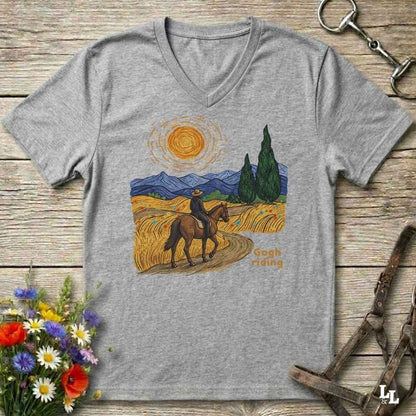 A heather gray V-neck t-shirt laid flat on a rustic wooden surface, featuring a colorful graphic of a cowboy riding a horse through a golden field under a bright sun. The shirt is styled with equestrian tack and a flower bouquet.