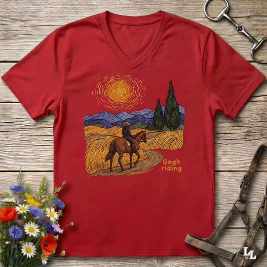 A red V-neck t-shirt laid flat on a rustic wooden surface, featuring a Gogh-style painting graphic of a cowboy riding a horse through a golden field under a swirling yellow sun, with the text 'Gogh riding'. The shirt is styled with horse tack, a halter, and a small bouquet of wildflowers.