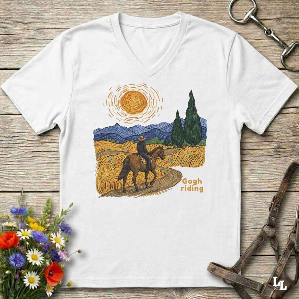A white V-neck t-shirt laid flat on a rustic wooden surface, featuring a colorful graphic of a cowboy riding a horse through a golden field under a bright sun, with the text 'Gogh riding'. The shirt is styled with equestrian props (reins and bridle) and a small bouquet of wildflowers.