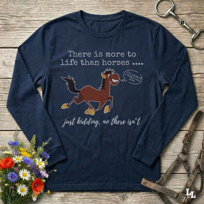 A navy blue long-sleeve graphic t-shirt laid flat on a rustic wooden surface, surrounded by equestrian-themed props including a halter, a horse bit, and a small bouquet of wildflowers. The shirt features a cartoon graphic of a brown horse with a speech bubble that reads 'I'll always be your friend' and text reading 'There is more to life than horses .