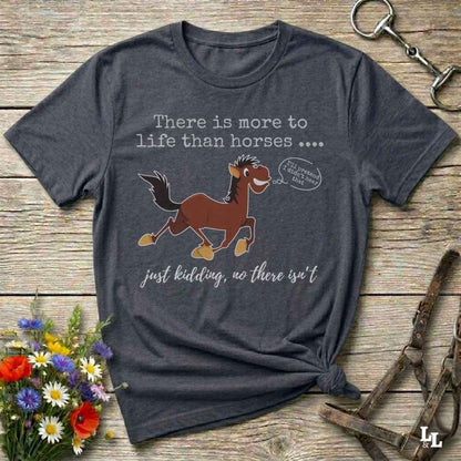 A dark heather gray graphic t-shirt laid flat on a rustic wooden background, styled with horse accessories and a small bouquet of wildflowers. The shirt features a cartoon illustration of a brown horse with a speech bubble t