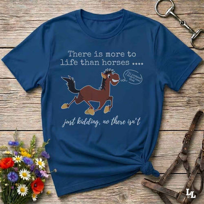 A navy blue graphic t-shirt laid flat on a rustic wooden surface, styled with horse tack and a small bouquet of flowers. The shirt features a cartoon illustration of a brown horse with a speech bubble that reads 'I'll show you how to ride!' and text that says 'There is more to life than horses .