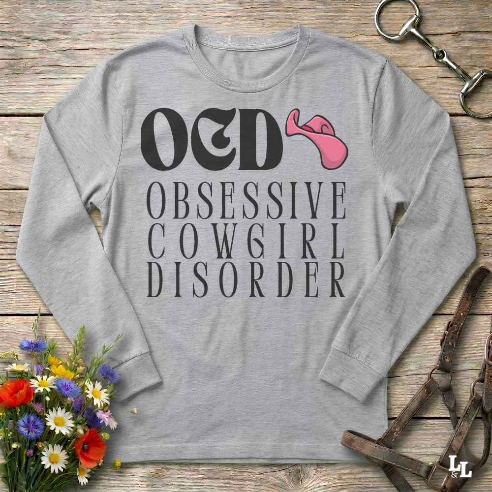 A light heather gray long-sleeve t-shirt with a graphic print reading 'O&D OBSESSIVE COWGIRL DISORDER' in black, featuring a pink cowgirl hat icon. The shirt is displayed in a flat-lay arrangement on a rustic wooden background with decorative equestrian and floral props.