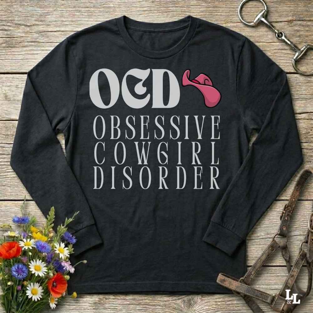 A black long-sleeve graphic t-shirt laid flat on a rustic wooden surface, featuring the text 'O&D OBSESSIVE COW6IR DISORDER' in white with a pink tongue-and-gag graphic. The shirt is styled with a horse bit in the top right corner and a small bouquet of colorful wildflowers in the bottom left corner.