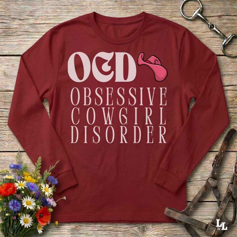 A  red long-sleeve graphic t-shirt laid flat on a rustic wooden surface, featuring the text 'O&D OBSESSIVE COW6IR DISORDER' in white with a pink tongue-and-gag graphic. The shirt is styled with a horse bit in the top right corner and a small bouquet of colorful wildflowers in the bottom left corner.