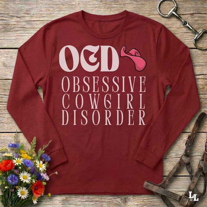 A  red long-sleeve graphic t-shirt laid flat on a rustic wooden surface, featuring the text 'O&D OBSESSIVE COW6IR DISORDER' in white with a pink tongue-and-gag graphic. The shirt is styled with a horse bit in the top right corner and a small bouquet of colorful wildflowers in the bottom left corner.