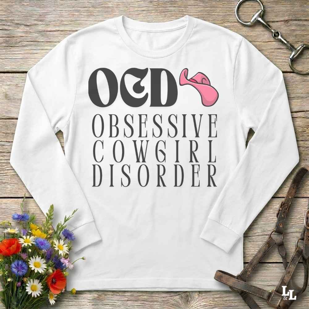 A white long-sleeve t-shirt with a graphic print reading 'O&D OBSESSIVE COWGIRL DISORDER' in black, accompanied by a pink cowboy hat illustration. The shirt is displayed flat on a rustic wooden background with equestrian-themed props including a halter, leather straps, and a small bouquet of flowers.