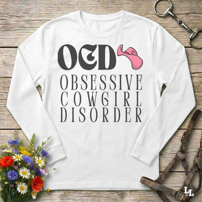 A white long-sleeve t-shirt with a graphic print reading 'O&D OBSESSIVE COWGIRL DISORDER' in black, accompanied by a pink cowboy hat illustration. The shirt is displayed flat on a rustic wooden background with equestrian-themed props including a halter, leather straps, and a small bouquet of flowers.