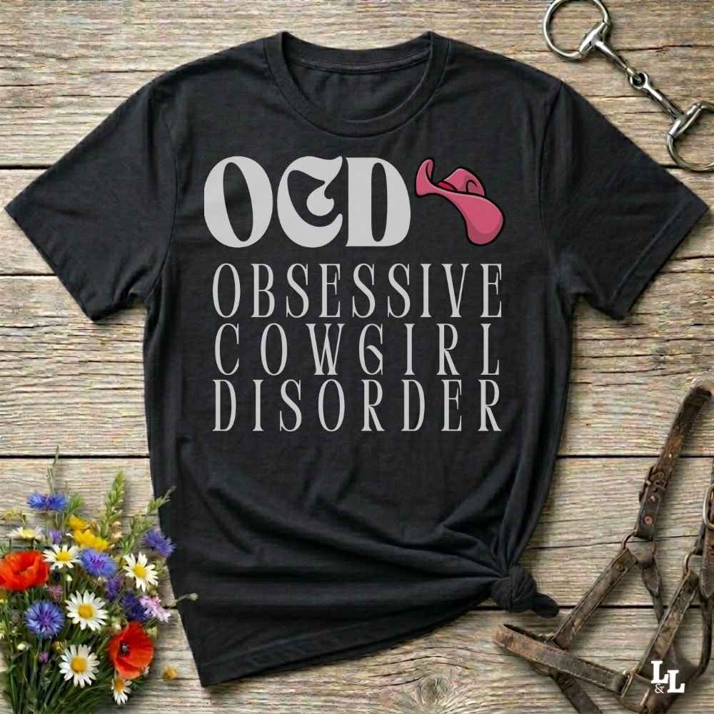 A black graphic t-shirt laid flat on a rustic wooden background, featuring the text 'O&D OBSESSIVE COWGIRL DISORDER' in white with a pink horn icon. The shirt is styled with a halter, flowers, and a horse bit.