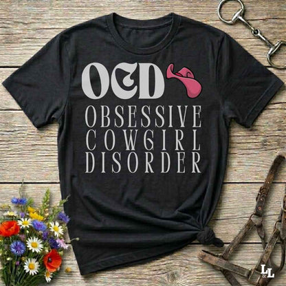 A black graphic t-shirt laid flat on a rustic wooden background, featuring the text 'O&D OBSESSIVE COWGIRL DISORDER' in white with a pink horn icon. The shirt is styled with a halter, flowers, and a horse bit.
