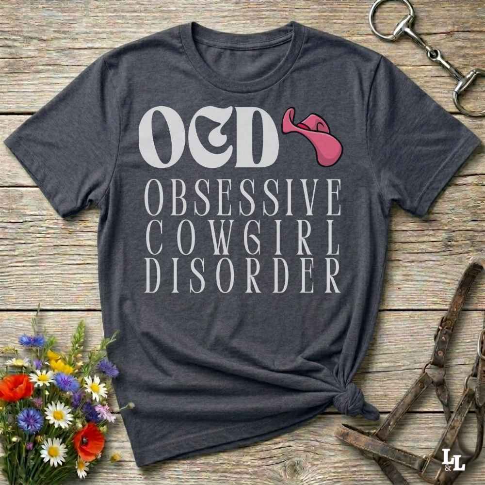 A dark heather gray graphic t-shirt laid flat on a rustic wooden surface, styled with a small bouquet of flowers, a horse bit, and a leather halter. The shirt features a graphic print that reads 'O&D OBSESSIVE COWGIRL DISORDER' in white, alongside a pink graphic resembling a stylized cowgirl hat or horn.