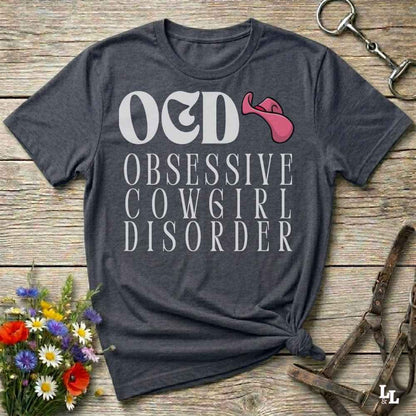 A dark heather gray graphic t-shirt laid flat on a rustic wooden surface, styled with a small bouquet of flowers, a horse bit, and a leather halter. The shirt features a graphic print that reads 'O&D OBSESSIVE COWGIRL DISORDER' in white, alongside a pink graphic resembling a stylized cowgirl hat or horn.
