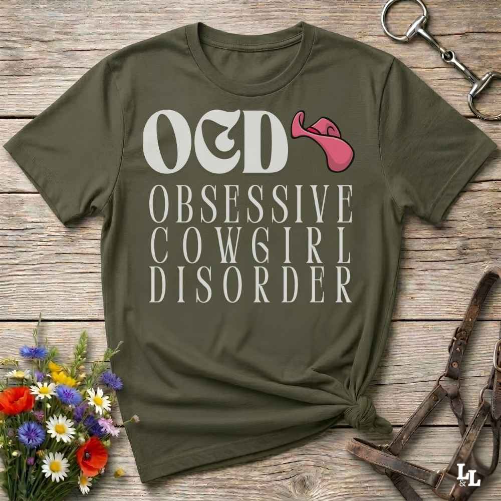 An olive green graphic t-shirt laid flat on a rustic wooden surface, styled with equestrian accessories and a small bouquet of flowers. The shirt features the text 'O&D OBSESSIVE COW6GIRL DISORDER' in white, with a pink horse bit graphic replacing the letter 'I' in 'O&D'.