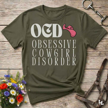An olive green graphic t-shirt laid flat on a rustic wooden surface, styled with equestrian accessories and a small bouquet of flowers. The shirt features the text 'O&D OBSESSIVE COW6GIRL DISORDER' in white, with a pink horse bit graphic replacing the letter 'I' in 'O&D'.