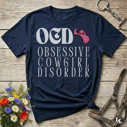 A navy blue graphic t-shirt laid flat on a rustic wooden surface, styled with equestrian accessories and a small bouquet of flowers. The shirt features the text 'O&D OBSESSIVE COWGIRL DISORDER' in white, alongside a pink horse bit graphic.