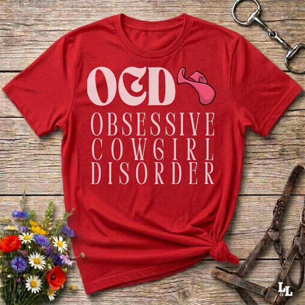 A red graphic t-shirt laid flat on a rustic wooden surface, styled with equestrian accessories and a small bouquet of flowers. The shirt features the text 'O&D OBSESSIVE COWGIRL DISORDER' in white, alongside a pink horse bit graphic.