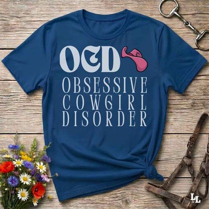 A navy blue graphic t-shirt laid flat on a rustic wooden surface, featuring the text 'O&D OBSESSIVE COW6IR DISORDER' in white with a pink tongue-and-gag graphic. The shirt is styled with a small bunch of wildflowers on the bottom left and brown leather tack on the right.
