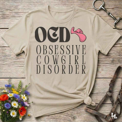 A beige graphic T-shirt laid flat on a rustic wood surface, with the text 'OCD OBSESSIVE COWGIRL DISORDER' in dark lettering and a pink cowgirl hat illustration. Decorative flowers and horse-riding accessories are placed around the shirt.