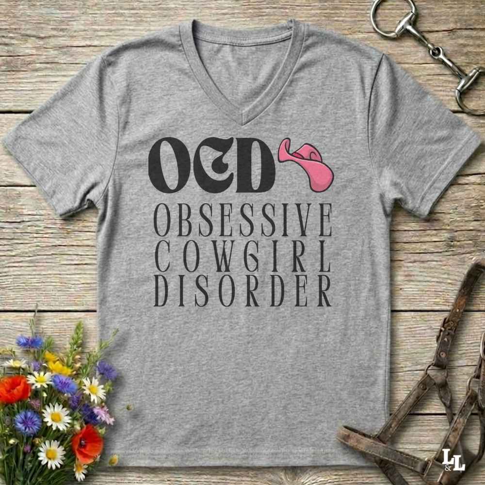 A grey V-neck t-shirt with a graphic print reading 'OED OBSESSIVE COWGIRL DISORDER' in white, featuring a pink cowboy hat accent, displayed on a rustic wooden background with equestrian and floral props.
