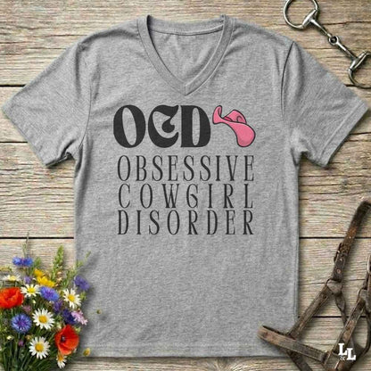 A grey V-neck t-shirt with a graphic print reading 'OED OBSESSIVE COWGIRL DISORDER' in white, featuring a pink cowboy hat accent, displayed on a rustic wooden background with equestrian and floral props.