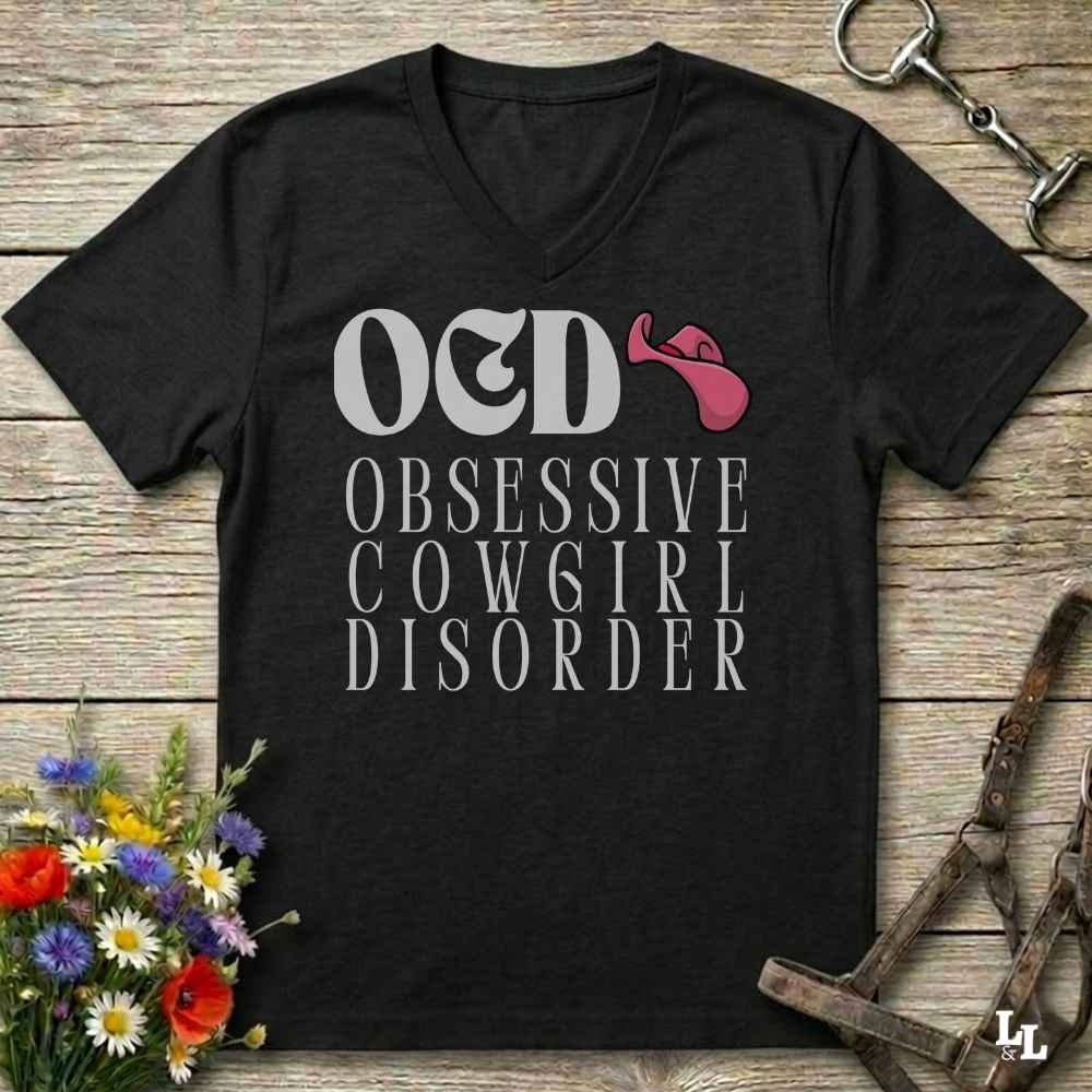 A black V-neck t-shirt with a graphic print reading 'OED OBSESSIVE COWGIRL DISORDER' in white, featuring a pink cowboy hat accent, displayed on a rustic wooden background with equestrian and floral props.