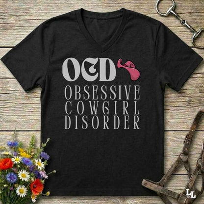A black V-neck t-shirt with a graphic print reading 'OED OBSESSIVE COWGIRL DISORDER' in white, featuring a pink cowboy hat accent, displayed on a rustic wooden background with equestrian and floral props.