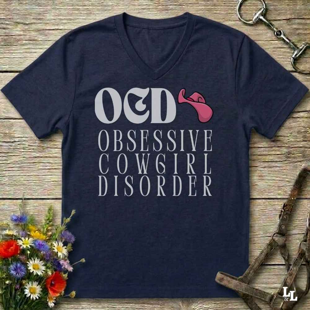 A blue V-neck t-shirt with a graphic print reading 'OED OBSESSIVE COWGIRL DISORDER' in white, featuring a pink cowboy hat accent, displayed on a rustic wooden background with equestrian and floral props.