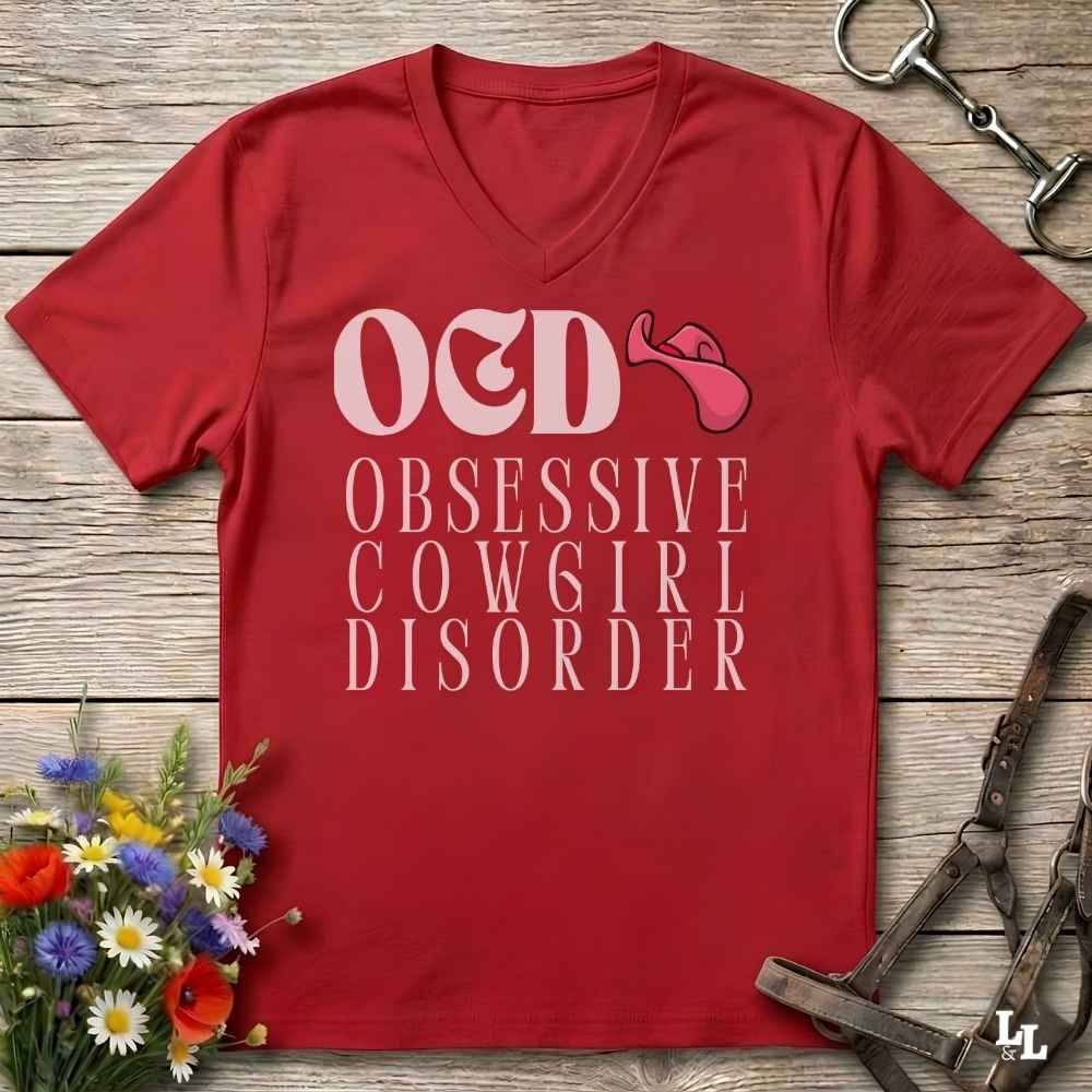 A red V-neck t-shirt laid flat on a rustic wooden surface, featuring the text 'OED OBSESSIVE COW6IRL DISORDER' in white with a pink cowboy hat graphic. The shirt is styled with equestrian gear and flowers around it.