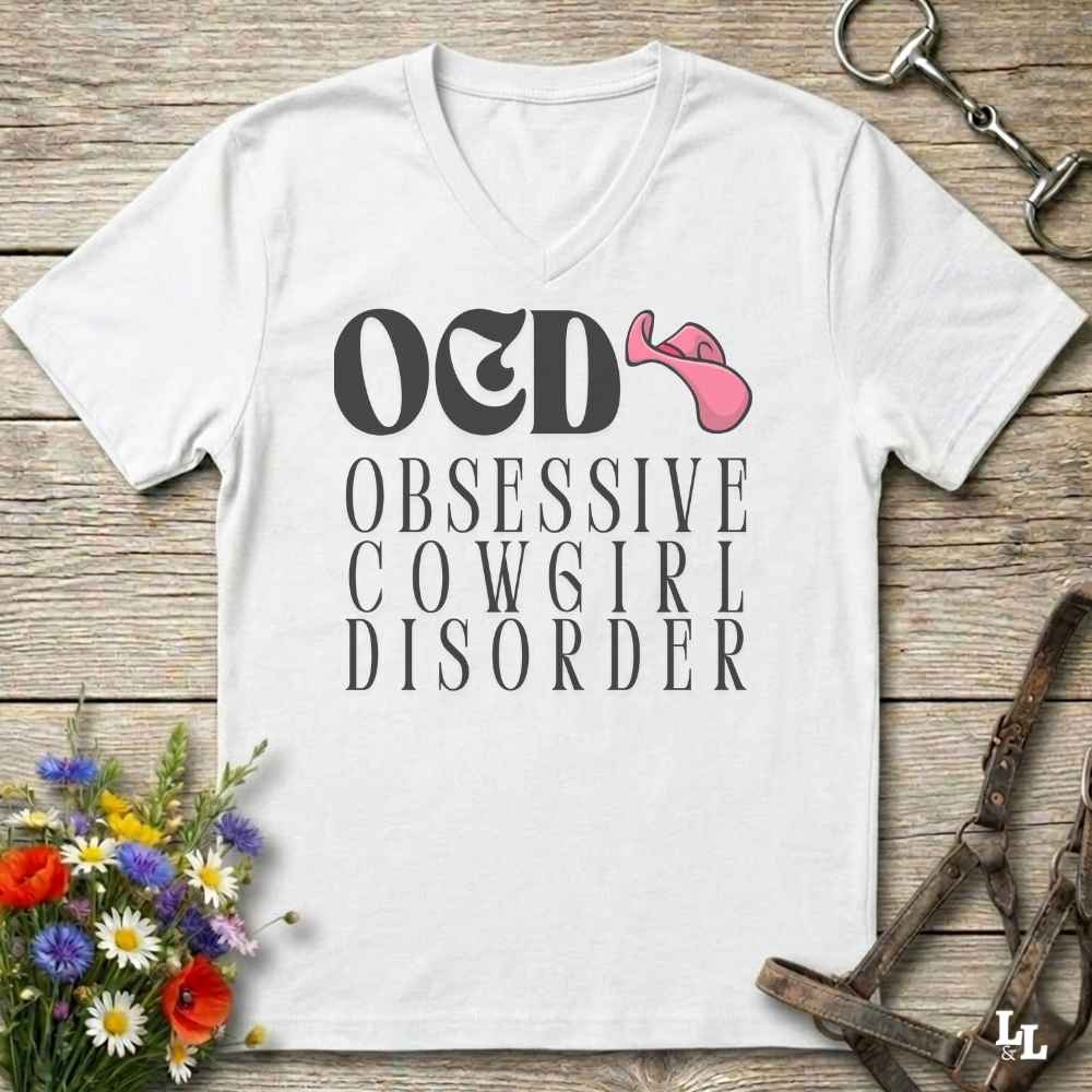 A white V-neck t-shirt laid flat on a rustic wooden surface, featuring the text 'O&D OBSESSIVE COWGIRL DISORDER' in black with a pink cowboy hat graphic. The shirt is styled with equestrian and floral props, including a halter, horse bit, and a small bouquet of wildflowers.