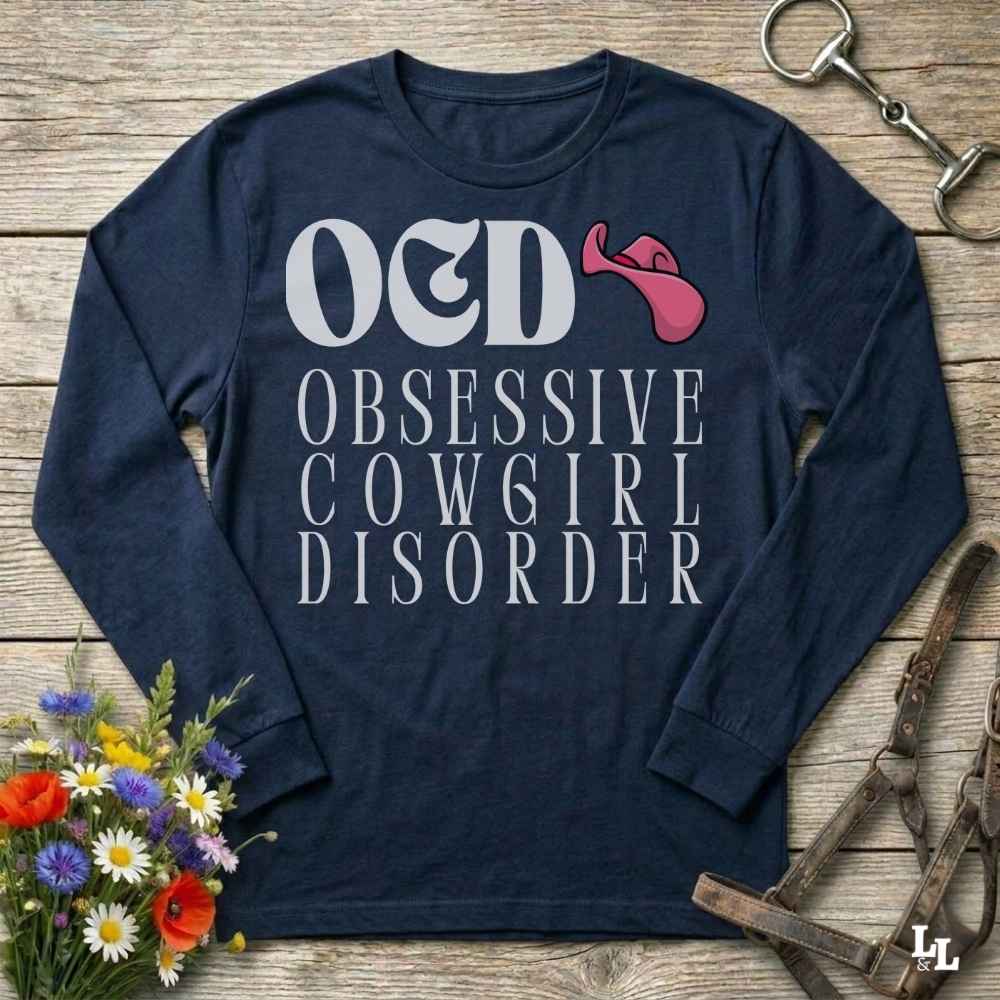 A navy blue long-sleeve graphic t-shirt laid flat on a rustic wooden surface, surrounded by cowgirl-themed props including a horse bit, leather bridle, and a small bouquet of wildflowers. The shirt features the text 'O&D OBSESSIVE COWGIRL DISORDER' in white, with a pink cowgirl hat graphic.
