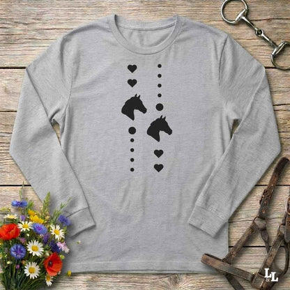 A light gray long-sleeve t-shirt with a black graphic of horse heads and hearts, laid flat on a rustic wooden surface with decorative flowers and leather tack around it.