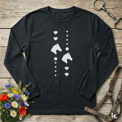 A black long-sleeve t-shirt with a white graphic of horse heads and hearts, laid flat on a rustic wooden surface with equestrian accessories and flowers around it.