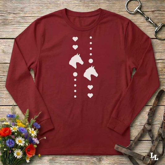 A long-sleeve crewneck t-shirt in maroon red, featuring a white graphic print of horse heads and hearts down the center. The shirt is laid flat on a rustic wooden background, styled with equestrian tack (a bit and bridle) and a small bouquet of colorful wildflowers.