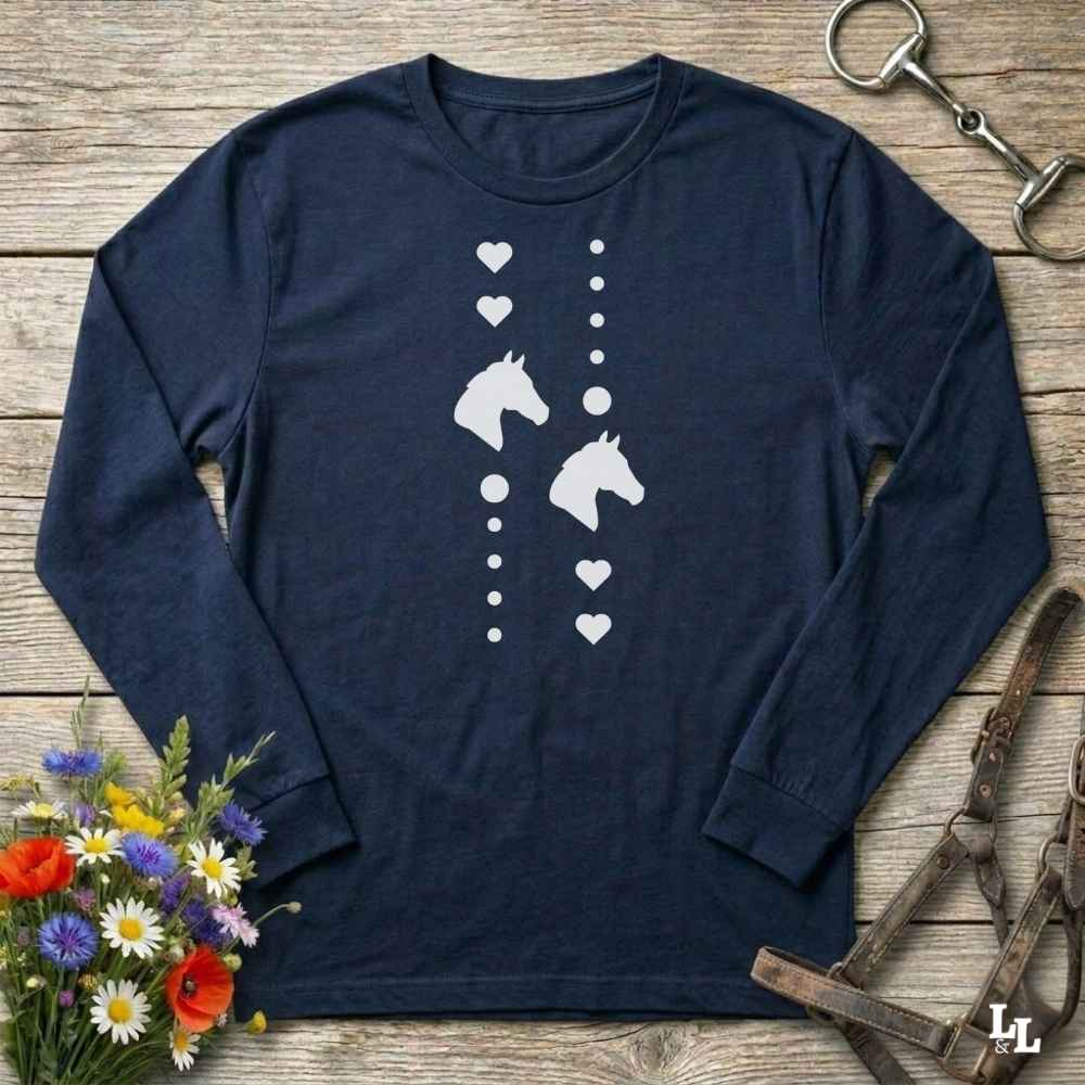 A navy blue long-sleeve t-shirt with a white graphic of horse heads and hearts, laid flat on a rustic wooden background with equestrian accessories and flowers.