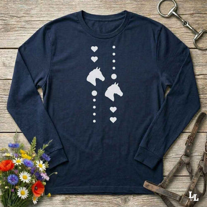 A navy blue long-sleeve t-shirt with a white graphic of horse heads and hearts, laid flat on a rustic wooden background with equestrian accessories and flowers.