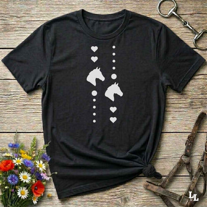 A black short-sleeve t-shirt with a white graphic print of horse heads and hearts, laid flat on a rustic wooden surface with equestrian-themed props.