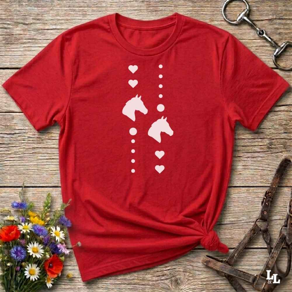 A red short-sleeve t-shirt with a white graphic print of horse heads and hearts, styled flat on a rustic wooden background with equestrian accessories and flowers.