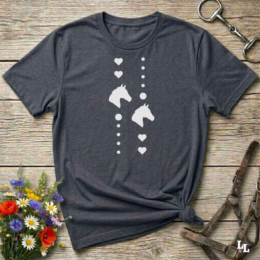 A dark charcoal gray t-shirt with a white graphic print of horse heads and hearts, styled in a flat-lay on a rustic wooden background with equestrian accessories and flowers.