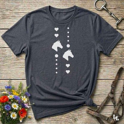 A dark charcoal gray t-shirt with a white graphic print of horse heads and hearts, styled in a flat-lay on a rustic wooden background with equestrian accessories and flowers.