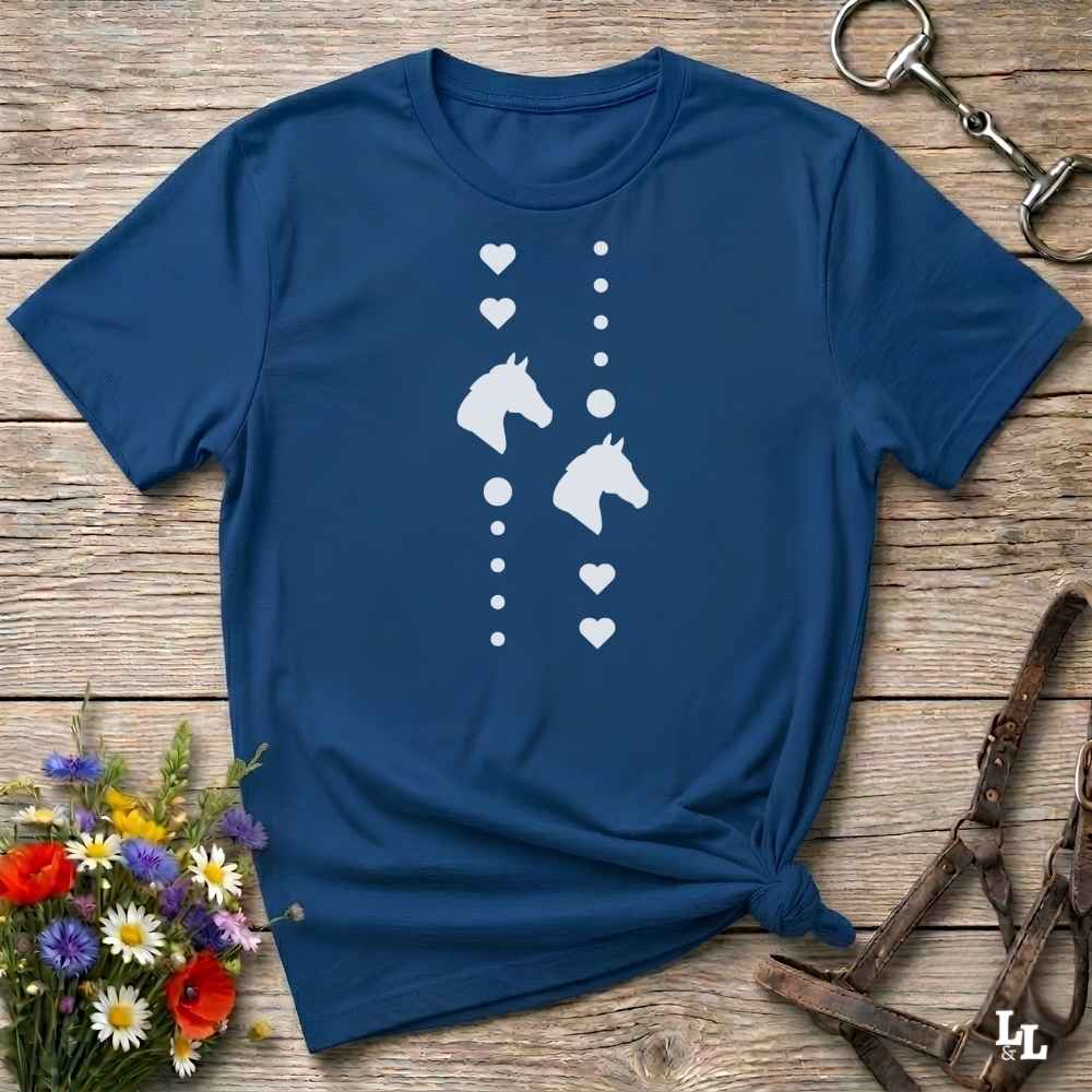 A navy blue short-sleeve t-shirt with a white graphic print of horse heads and hearts, styled in a flat-lay on a rustic wooden background with equestrian accessories and flowers.