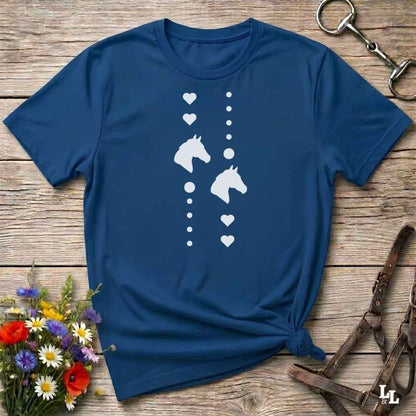 A navy blue short-sleeve t-shirt with a white graphic print of horse heads and hearts, styled in a flat-lay on a rustic wooden background with equestrian accessories and flowers.
