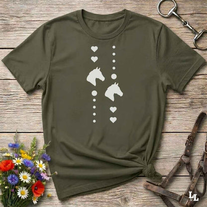 An olive green short-sleeve t-shirt with a white graphic print of horse heads and hearts, styled in a flat-lay on a rustic wooden background with equestrian accessories and flowers.