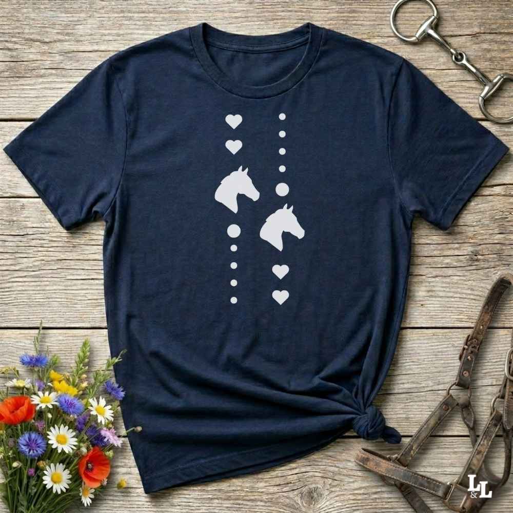 A navy blue short-sleeve t-shirt with a white graphic of two horse heads and hearts, laid flat on a rustic wooden background with equestrian accessories and flowers.