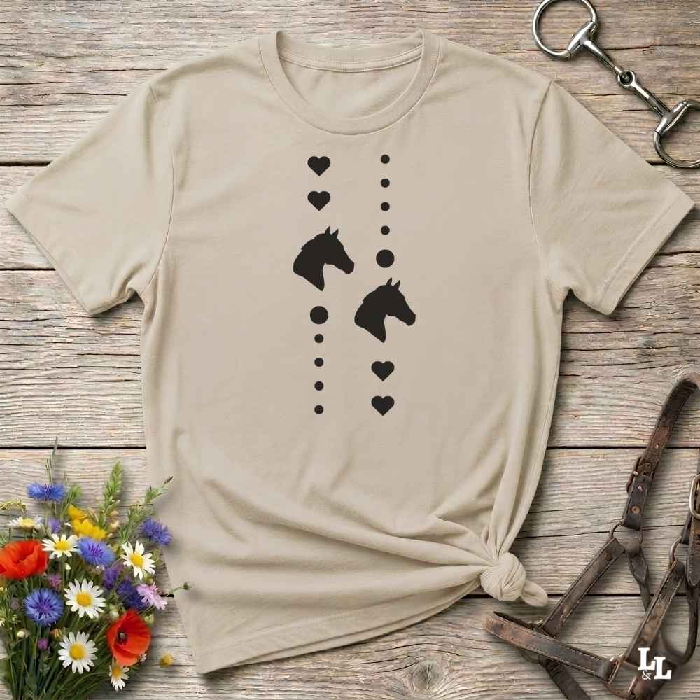 A beige short-sleeve t-shirt with a black graphic of horse heads and hearts, styled in a flat-lay on a rustic wooden background with equestrian accessories and flowers.