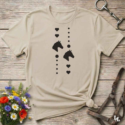 A beige short-sleeve t-shirt with a black graphic of horse heads and hearts, styled in a flat-lay on a rustic wooden background with equestrian accessories and flowers.