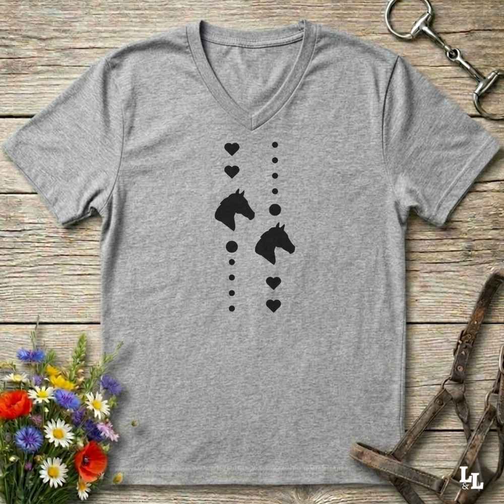A heather gray V-neck t-shirt with a black graphic of horse heads and hearts, laid flat on a rustic wooden surface with equestrian accessories and flowers around it.