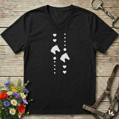 A black V-neck t-shirt with a white graphic print of horse heads, hearts, and dots, laid flat on a rustic wooden background with equestrian accessories and flowers.