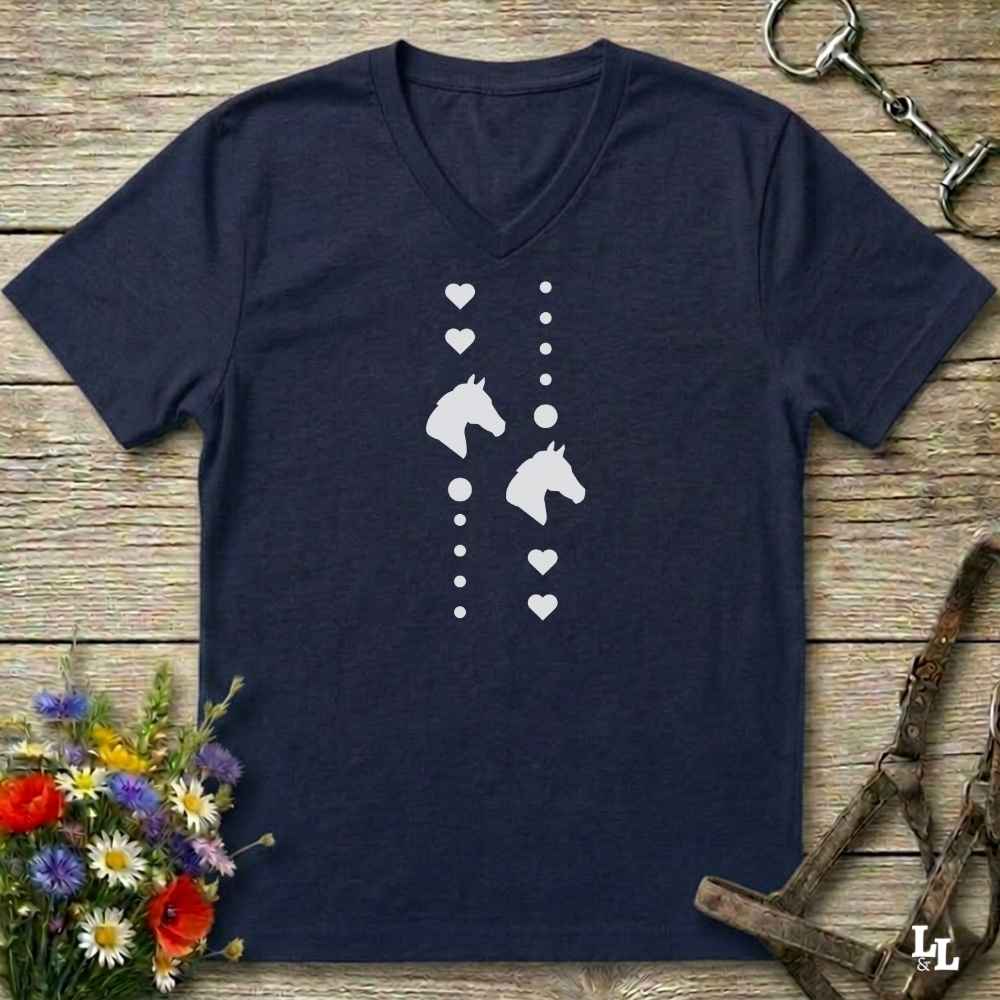 A navy blue V-neck t-shirt with a white graphic print of horse heads, hearts, and dots, laid flat on a rustic wooden background with equestrian accessories and flowers.